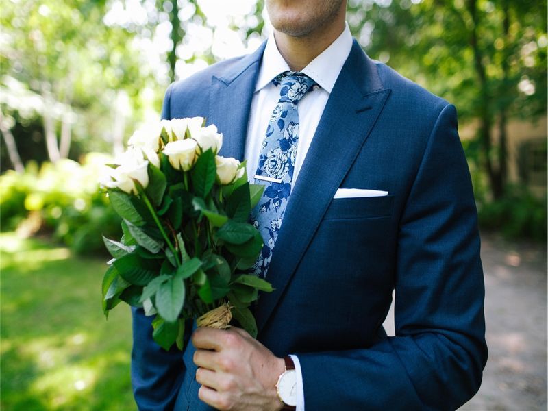 A man stands in a blue suit and tie with white flowers in his hand - Ideas for a prom-themed birthday party - Image